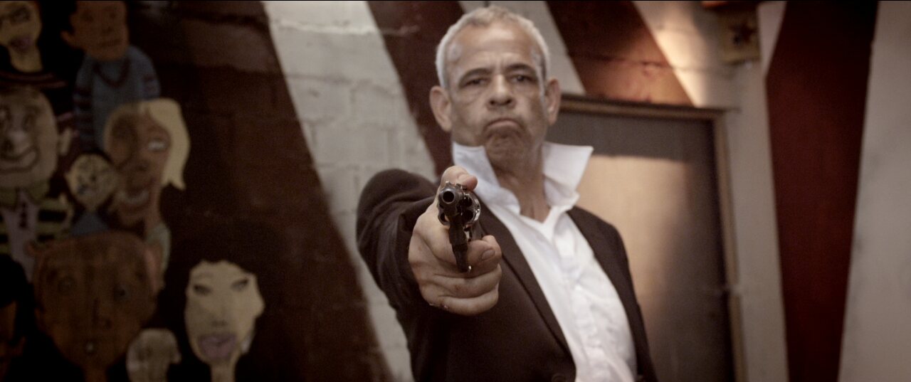Film still: a man aiming a revolver at camera in front of painted faces