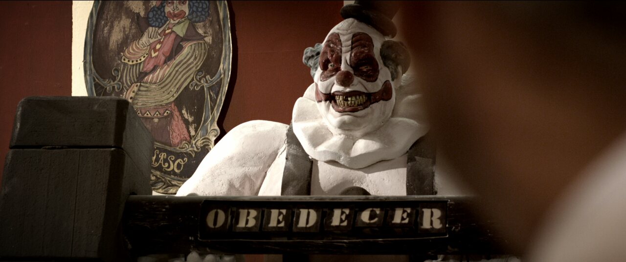 Film still: a clown figure above a plaque reading OBEDECER